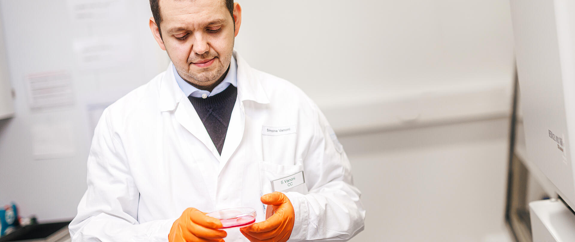 Ein Wissenschaftler im weißen Laborkittel und mit orangefarbenen Handschuhen betrachtet konzentriert eine Petrischale mit einer rötlichen Substanz. Er steht in einem hellen Labor vor medizinischen Geräten und Arbeitsstationen.
